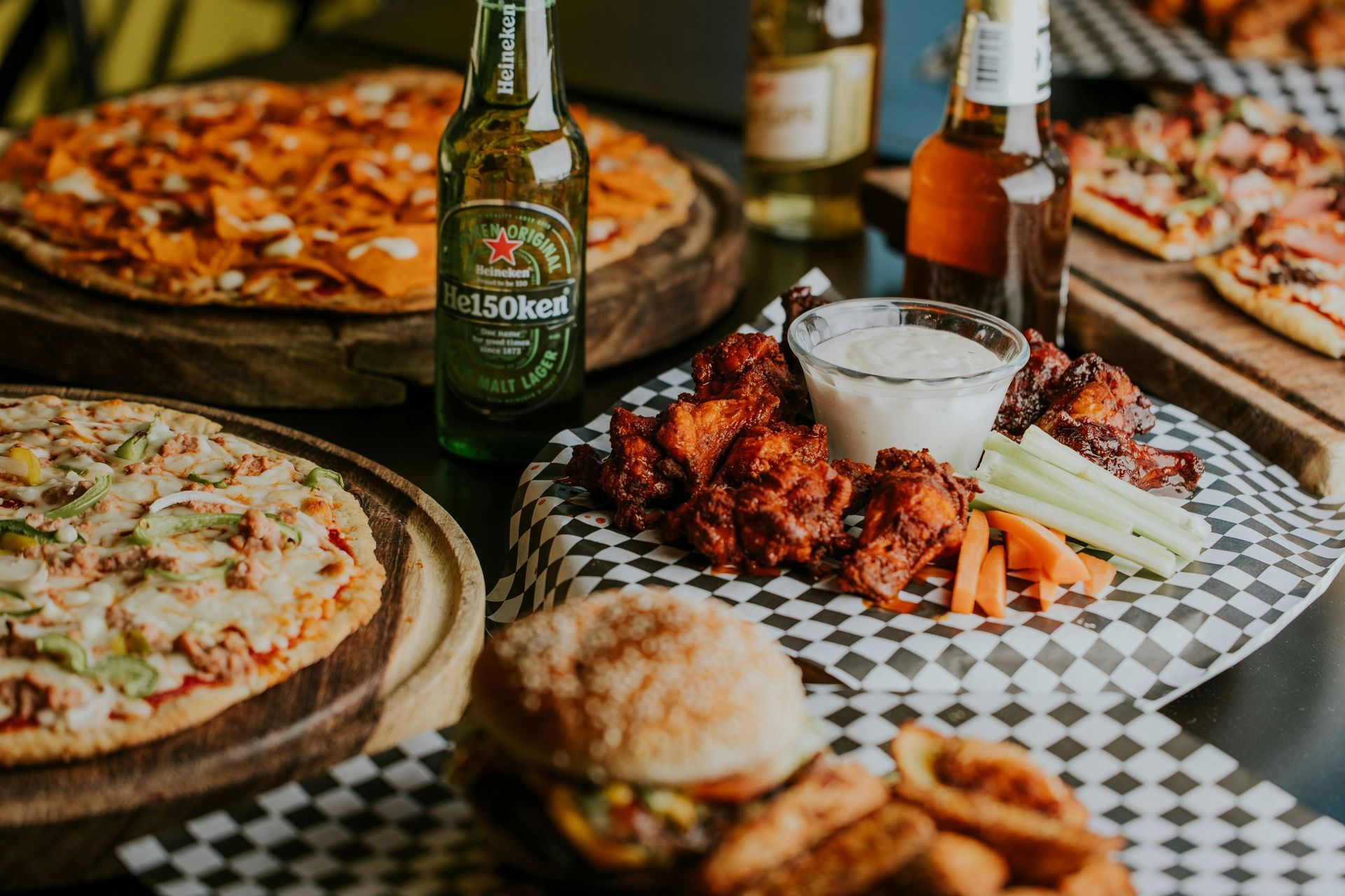 A table topped with pizza , wings , a hamburger , and a bottle of beer.