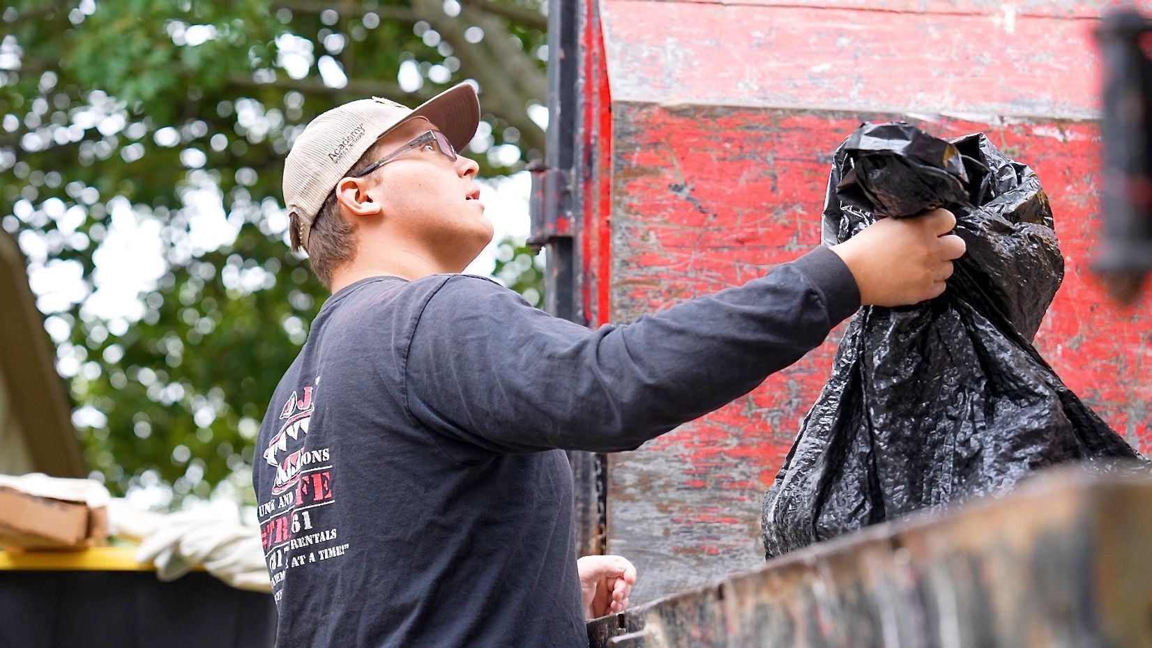 Person in a cap throwing a black trash bag into a red dumpster, outdoors.
