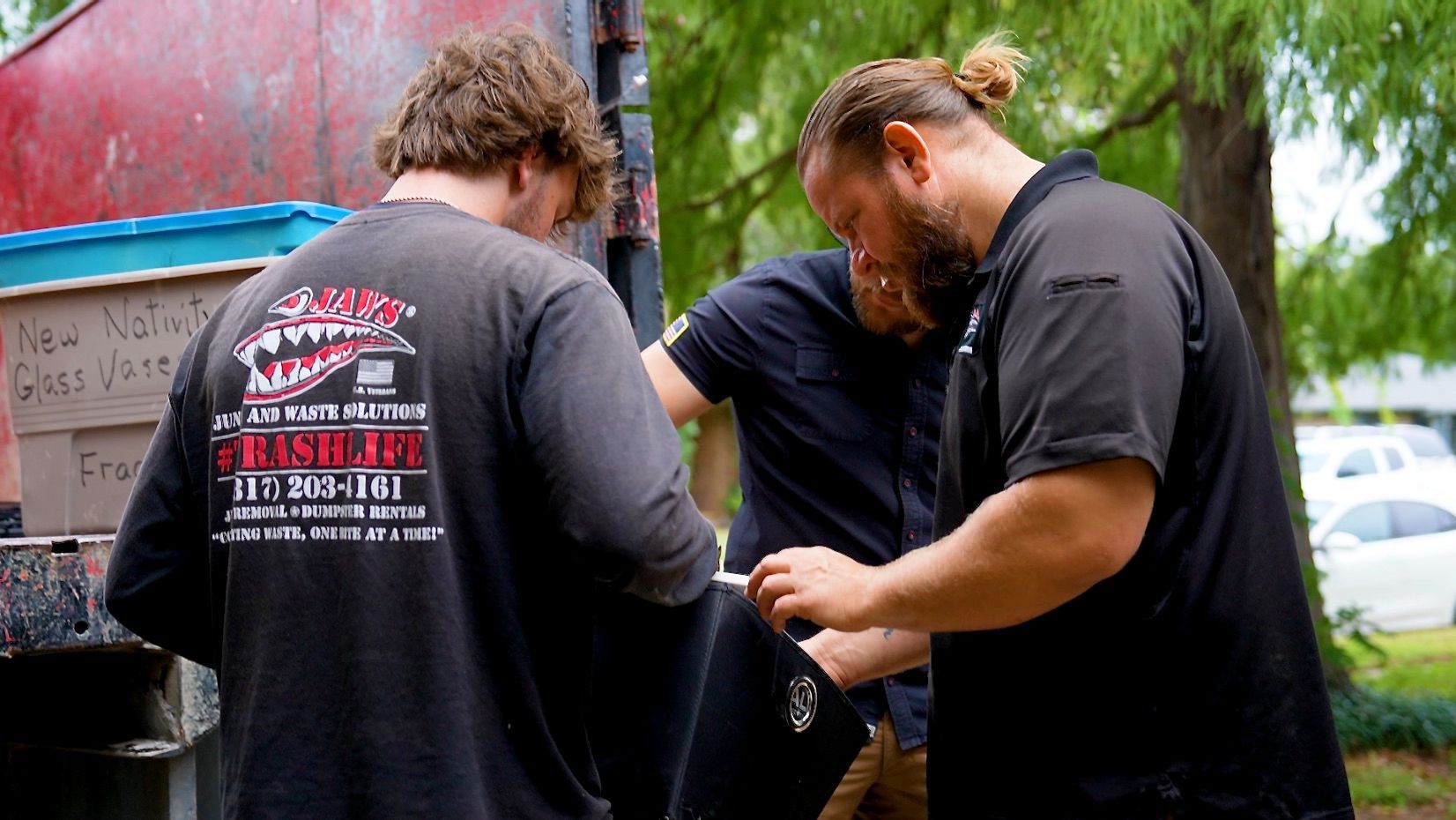 Three men examining something, likely working on a truck, outdoors. Two wear black shirts, one with a logo.