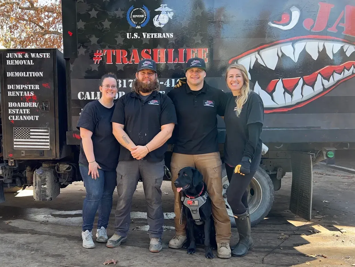 Four people and a dog pose in front of a trash truck.  The people are smiling, with the truck in the background.