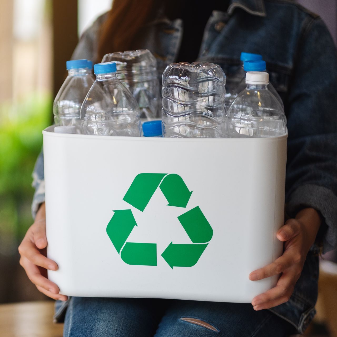 A woman is holding a recycling bin filled with plastic bottles.