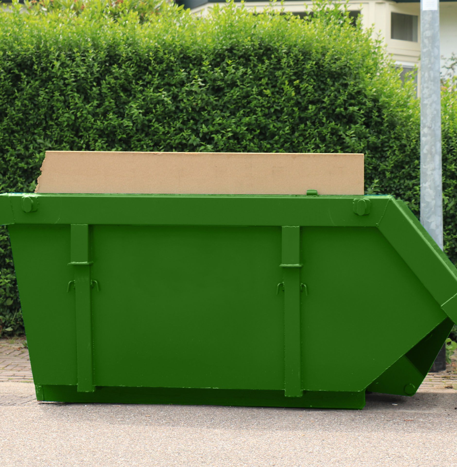 A green skip bin on the side of the road next to a hedge.