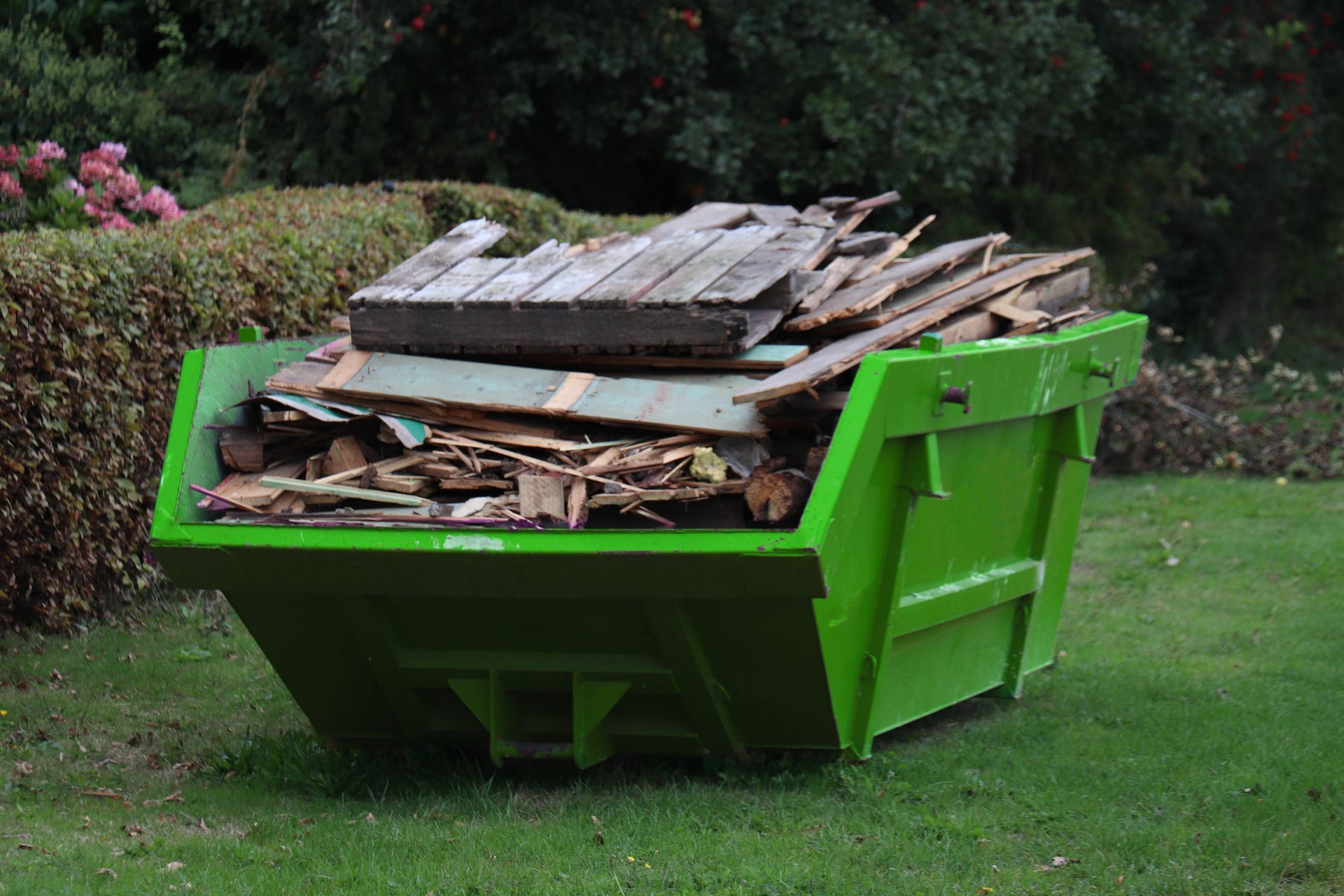 A green skip bin filled with wood is sitting on top of a lush green lawn.