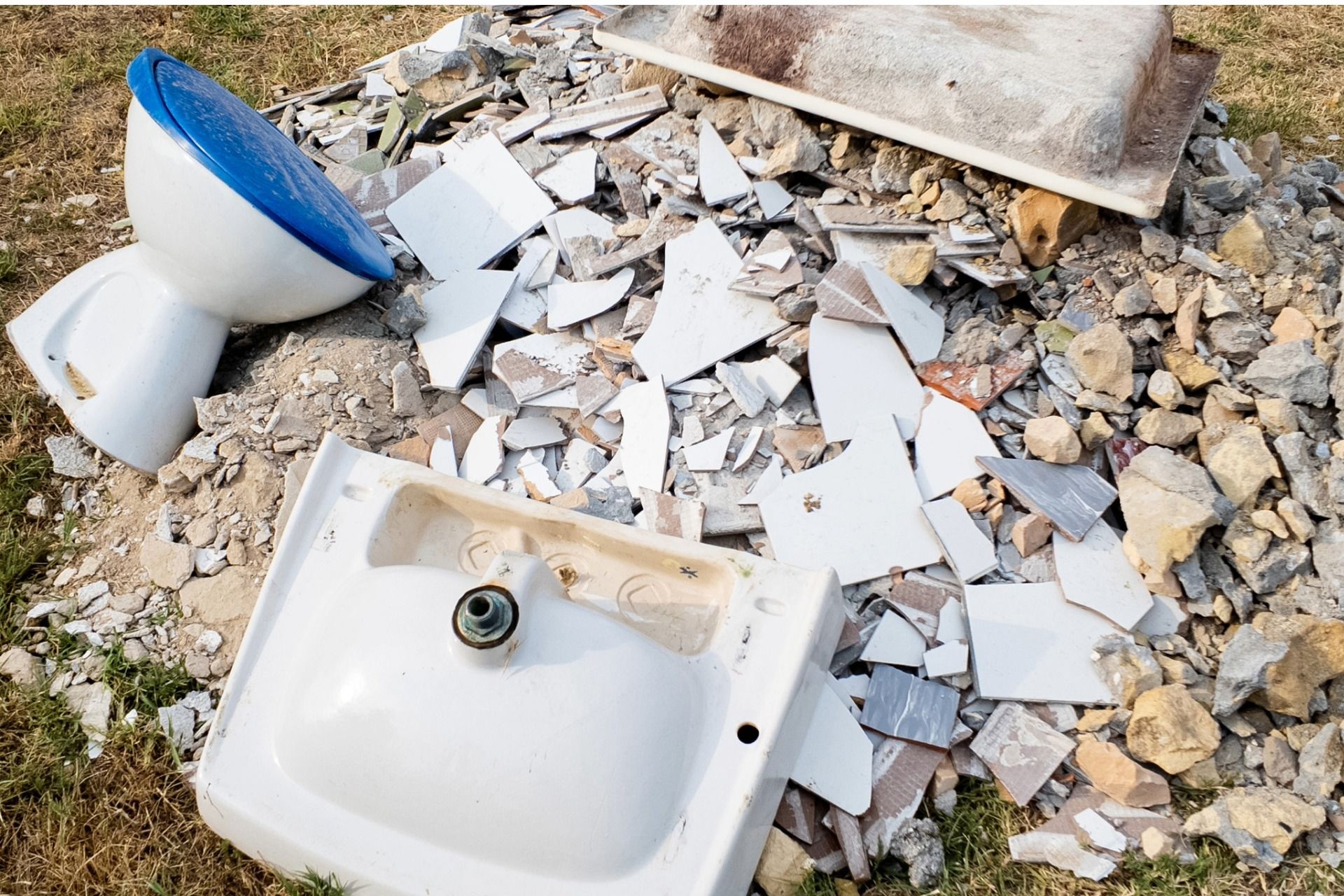 Pile of broken bathroom fixtures: toilet, sink, tub, and tile rubble outdoors.
