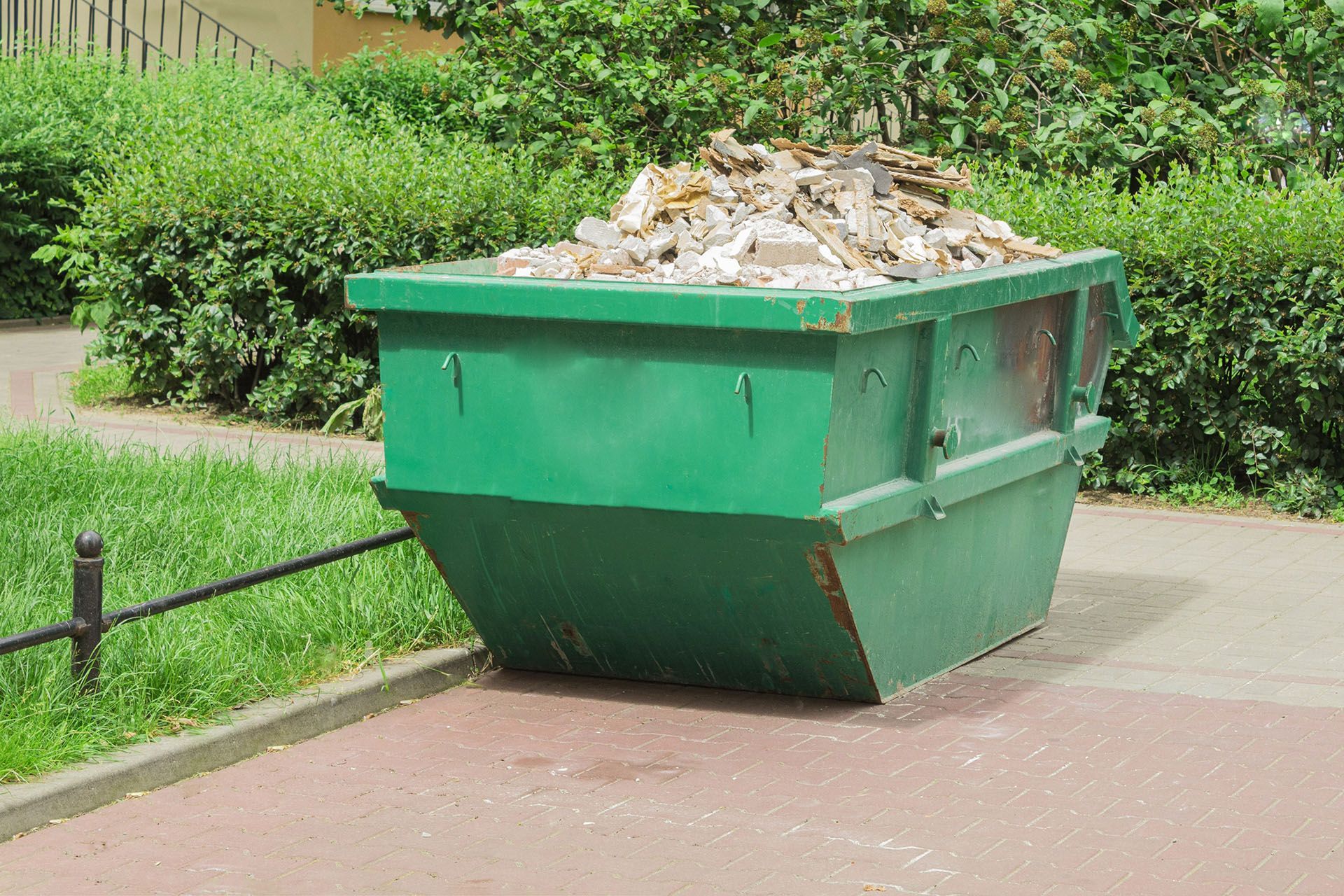 Green dumpster overflowing with debris on a red brick sidewalk near green bushes.