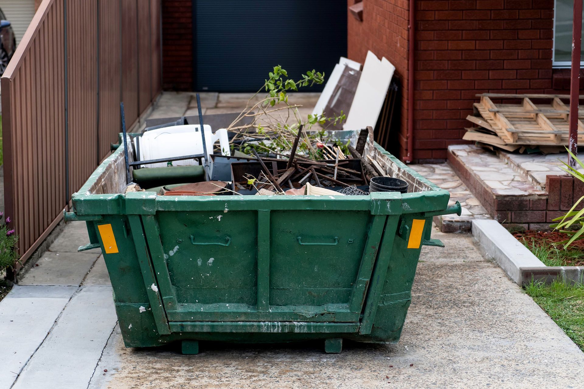Green dumpster overflowing with debris on a concrete driveway next to a brick building.