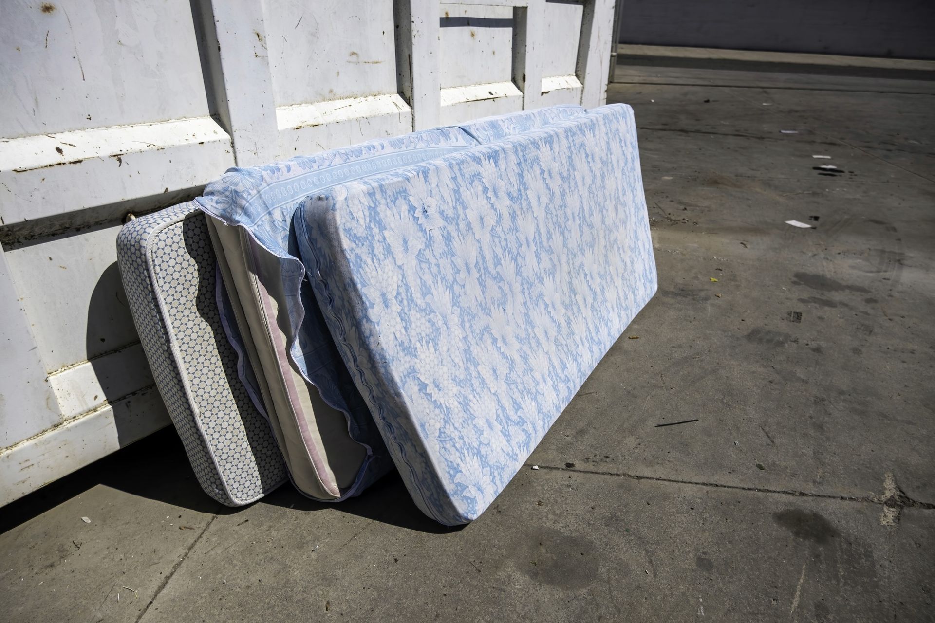 Three discarded mattresses lean against a dumpster, outdoors on a concrete surface.