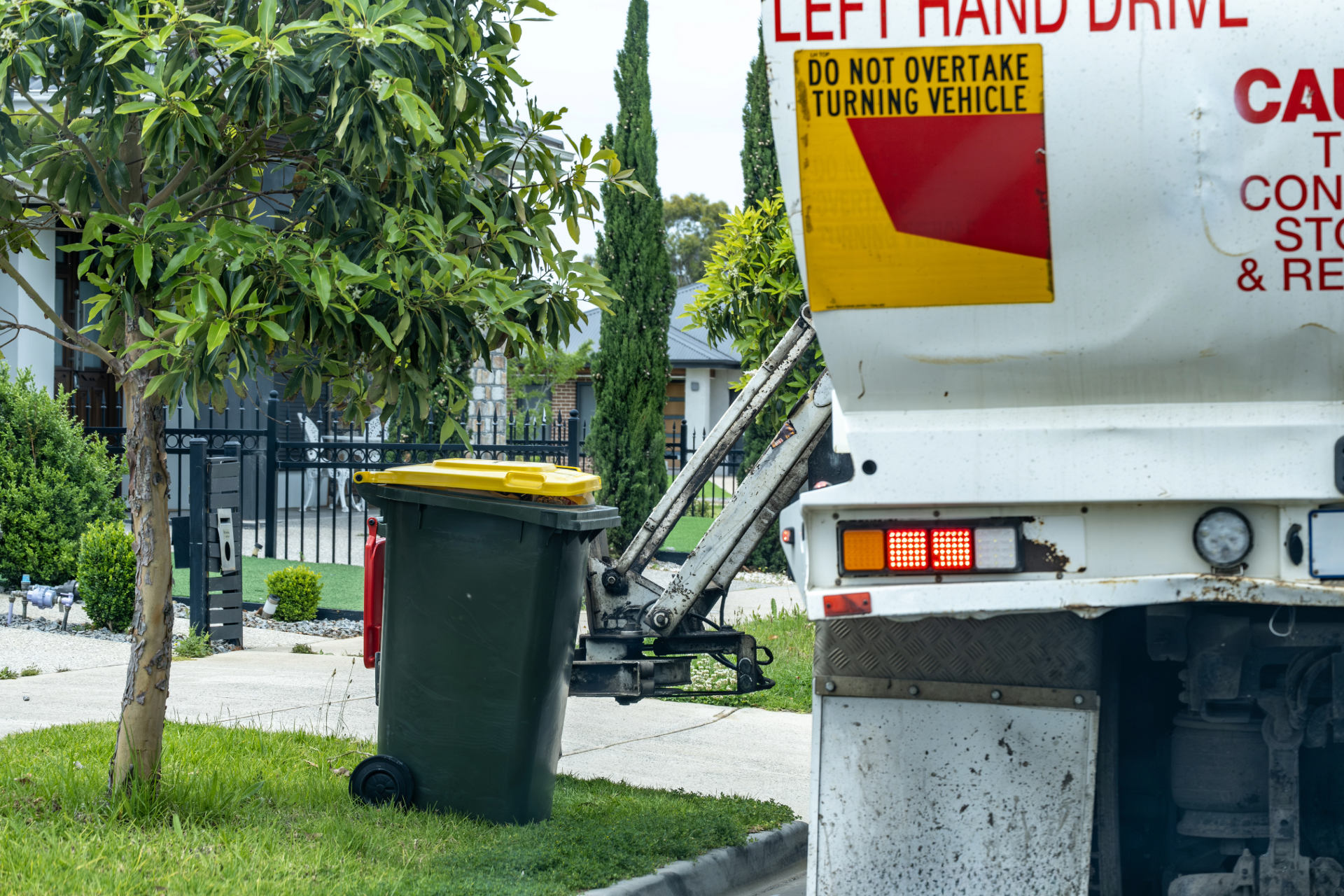 Garbage worker in orange vest and hard hat, loading yellow bin into green truck on a sunny street.