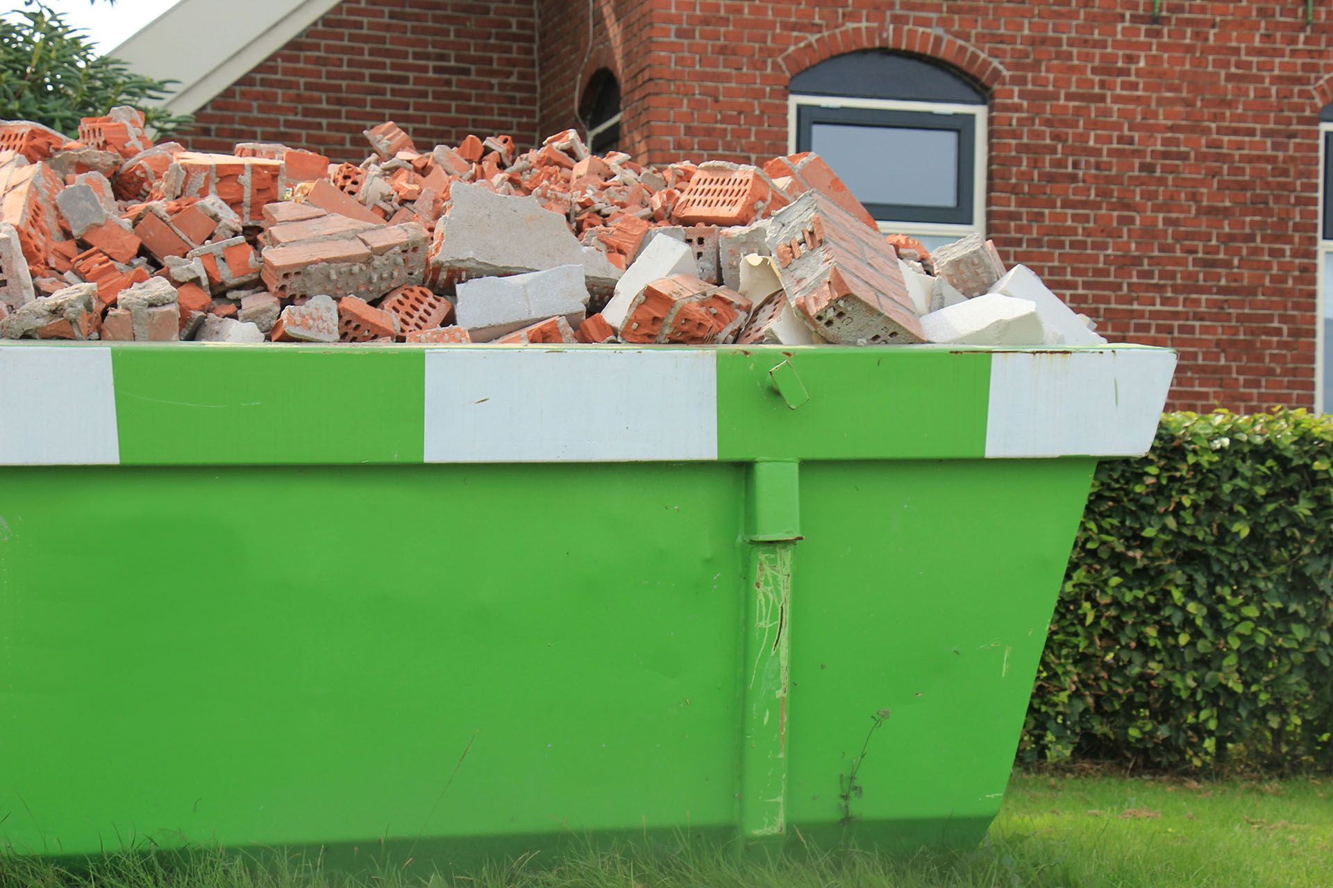 Green dumpster filled with brick and construction debris in front of a red brick building.