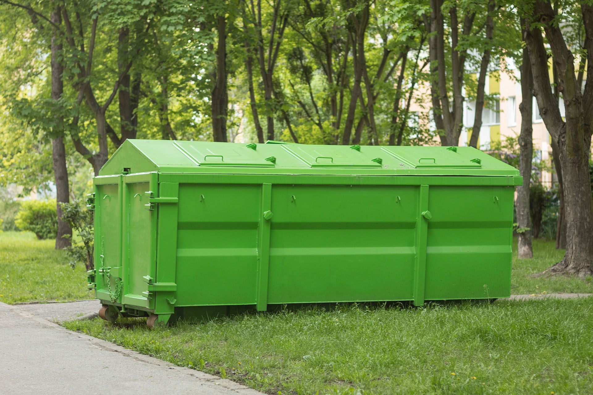 Green dumpster on a grassy area, next to a sidewalk, with trees in the background.