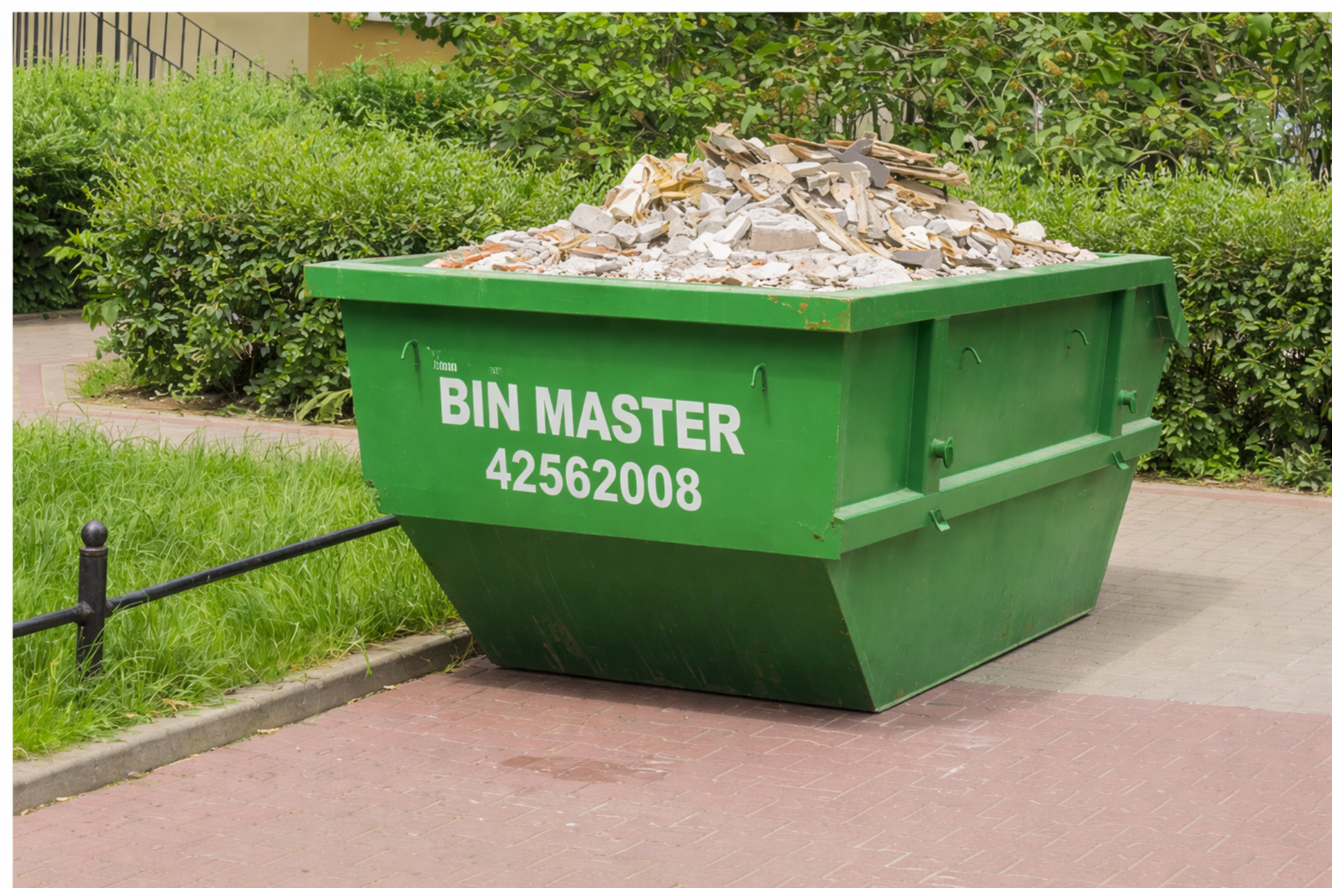 Green dumpster overflowing with debris on a red brick sidewalk near green bushes.