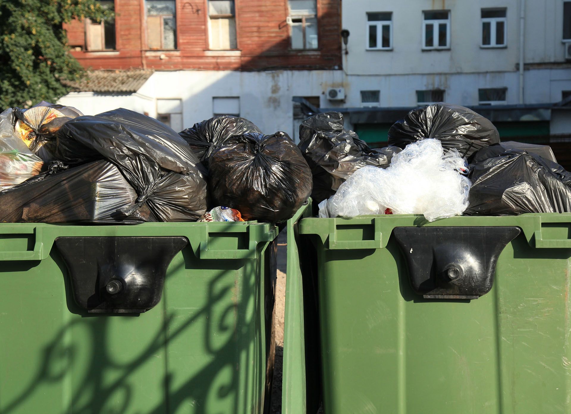 Green trash bins overflowing with black garbage bags against a blurred building background.
