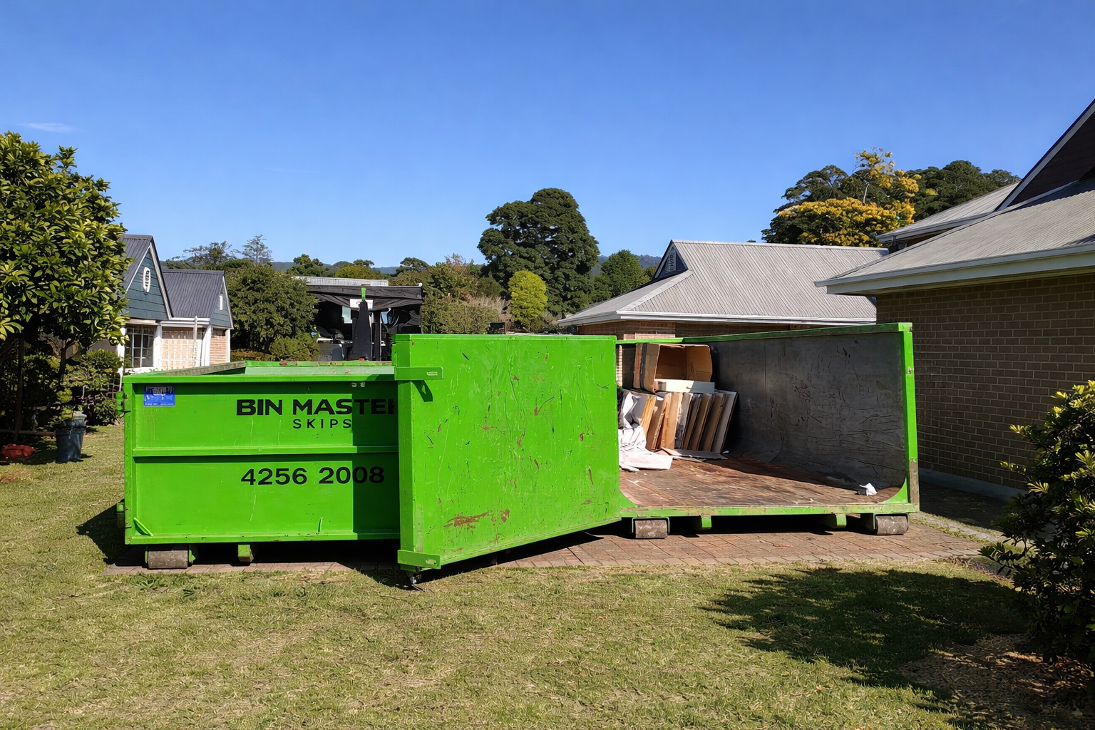 Green dumpster overflowing with debris on a concrete driveway next to a brick building.