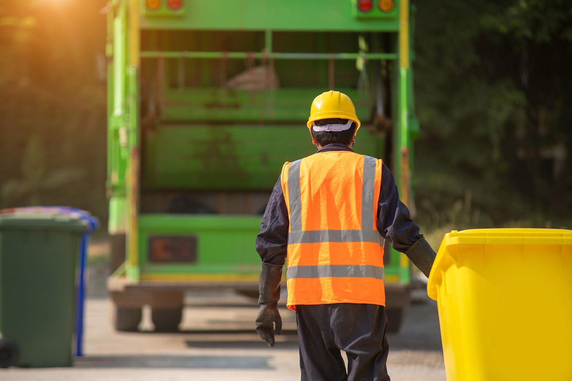 Garbage worker in orange vest and hard hat, loading yellow bin into green truck on a sunny street.