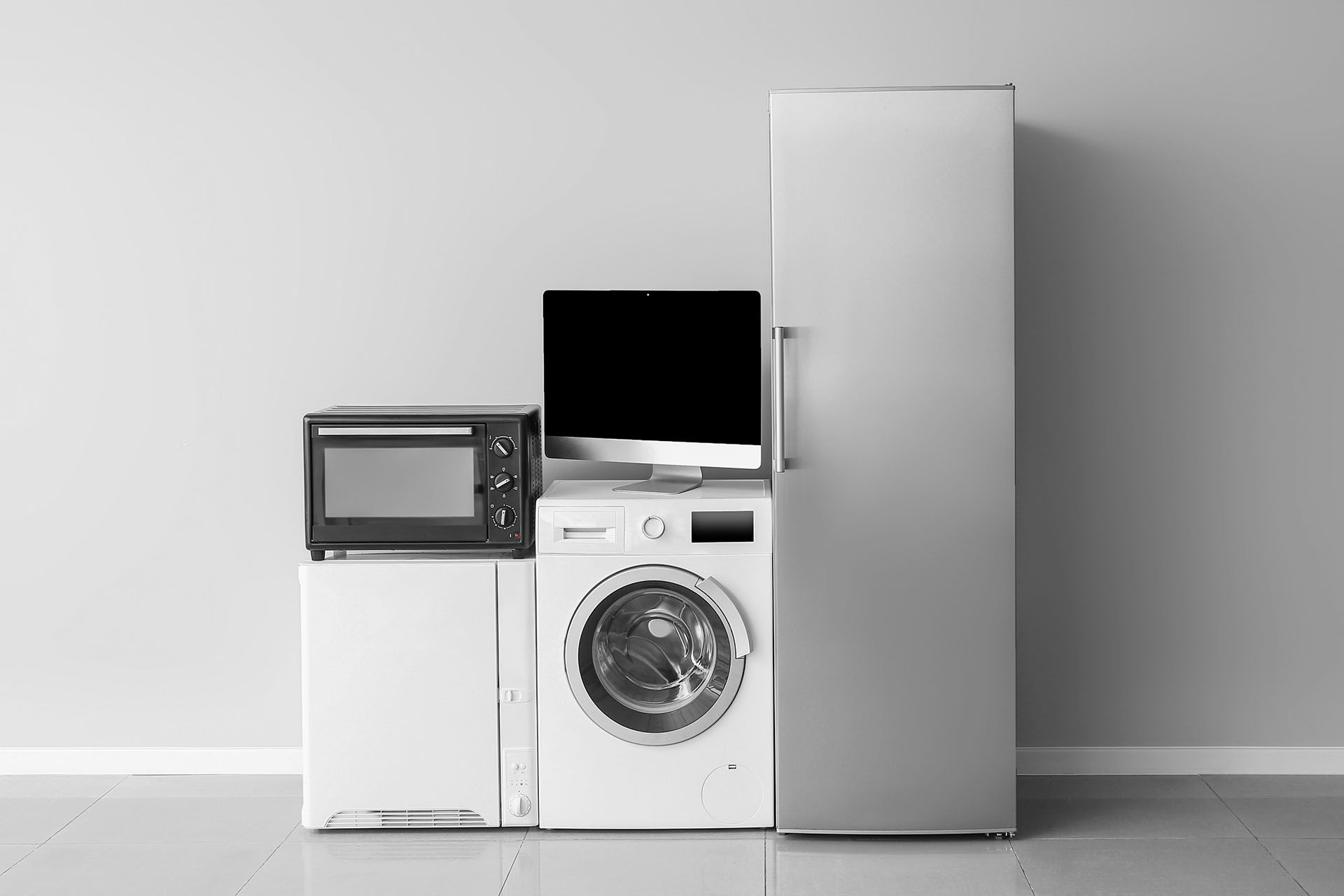 Black and white photo of appliances: microwave, TV, washing machine, and refrigerator against a plain wall.
