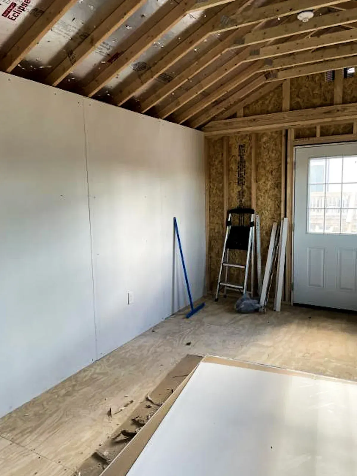 Interior of a room under construction with white walls, exposed rafters, door, and a ladder.
