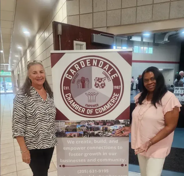 Two women stand by a Gardendale Chamber of Commerce banner. One wears zebra print, the other, pink. Indoors.