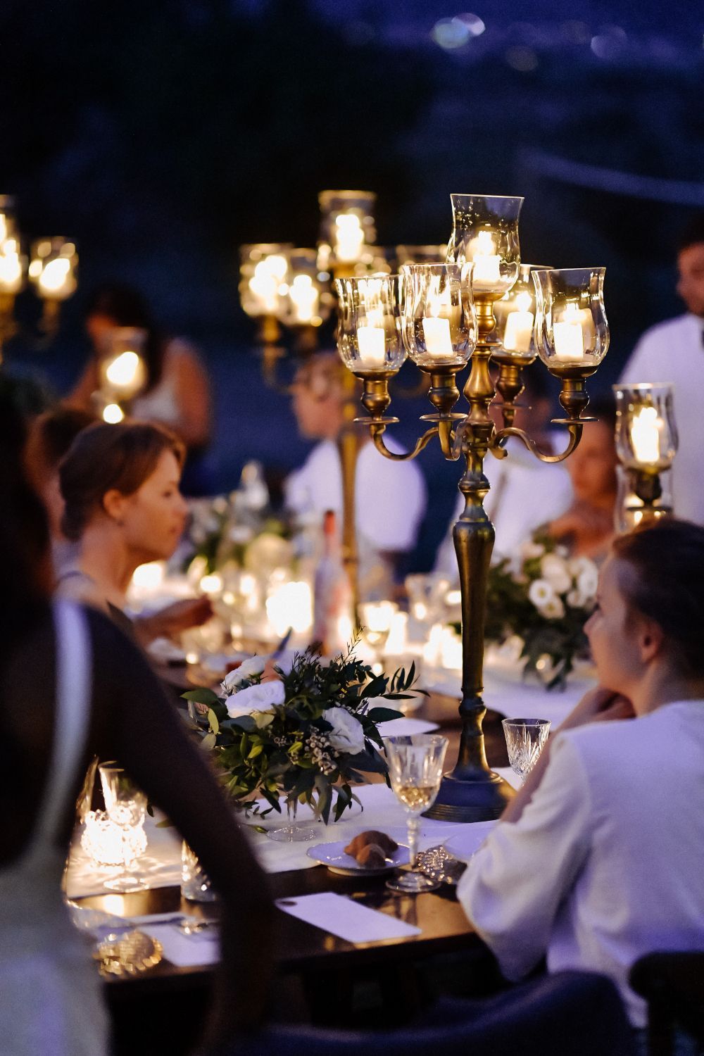 Candlelit dinner party outdoors at night; people seated at a table with ornate candelabras and floral centerpieces.