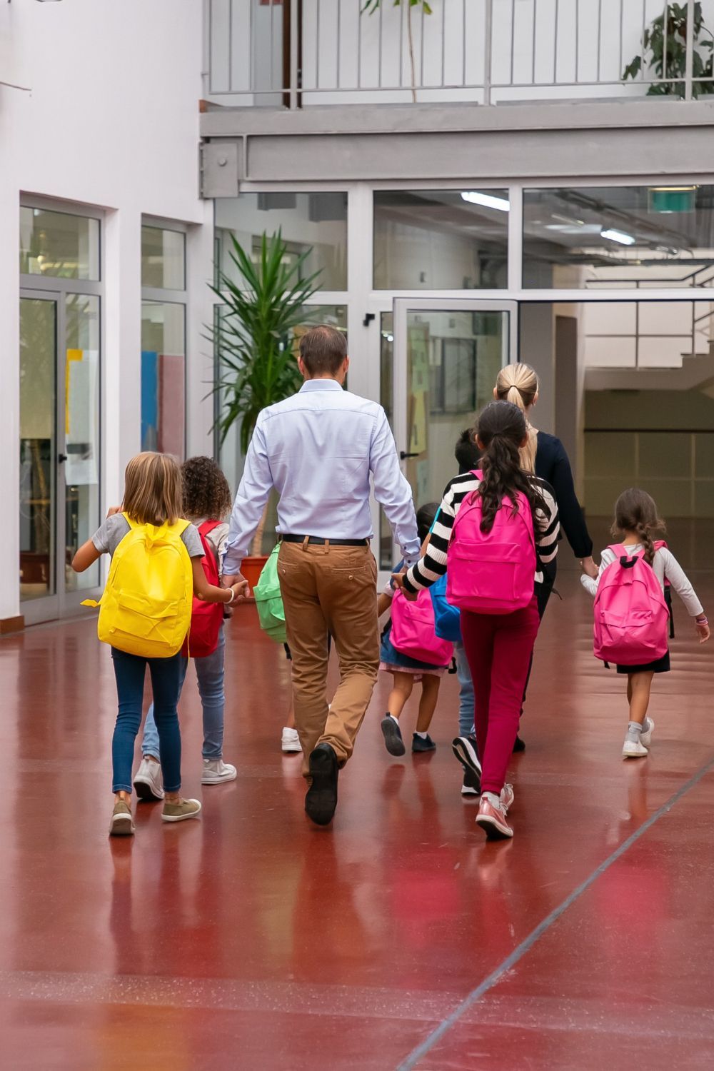 Group of children and adults walking inside a building, holding hands, with backpacks.