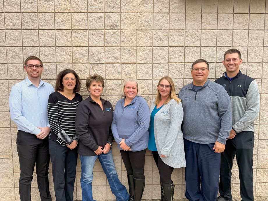 Woman in a black suit smiles while talking on a landline phone in an office setting.