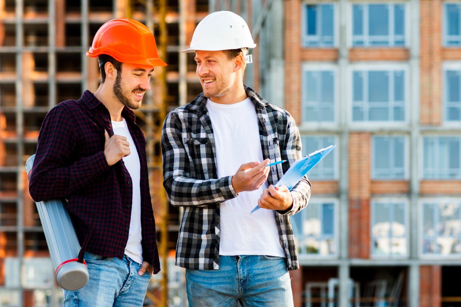 Two construction workers discussing plans at a building site; one with orange hard hat, one with white.