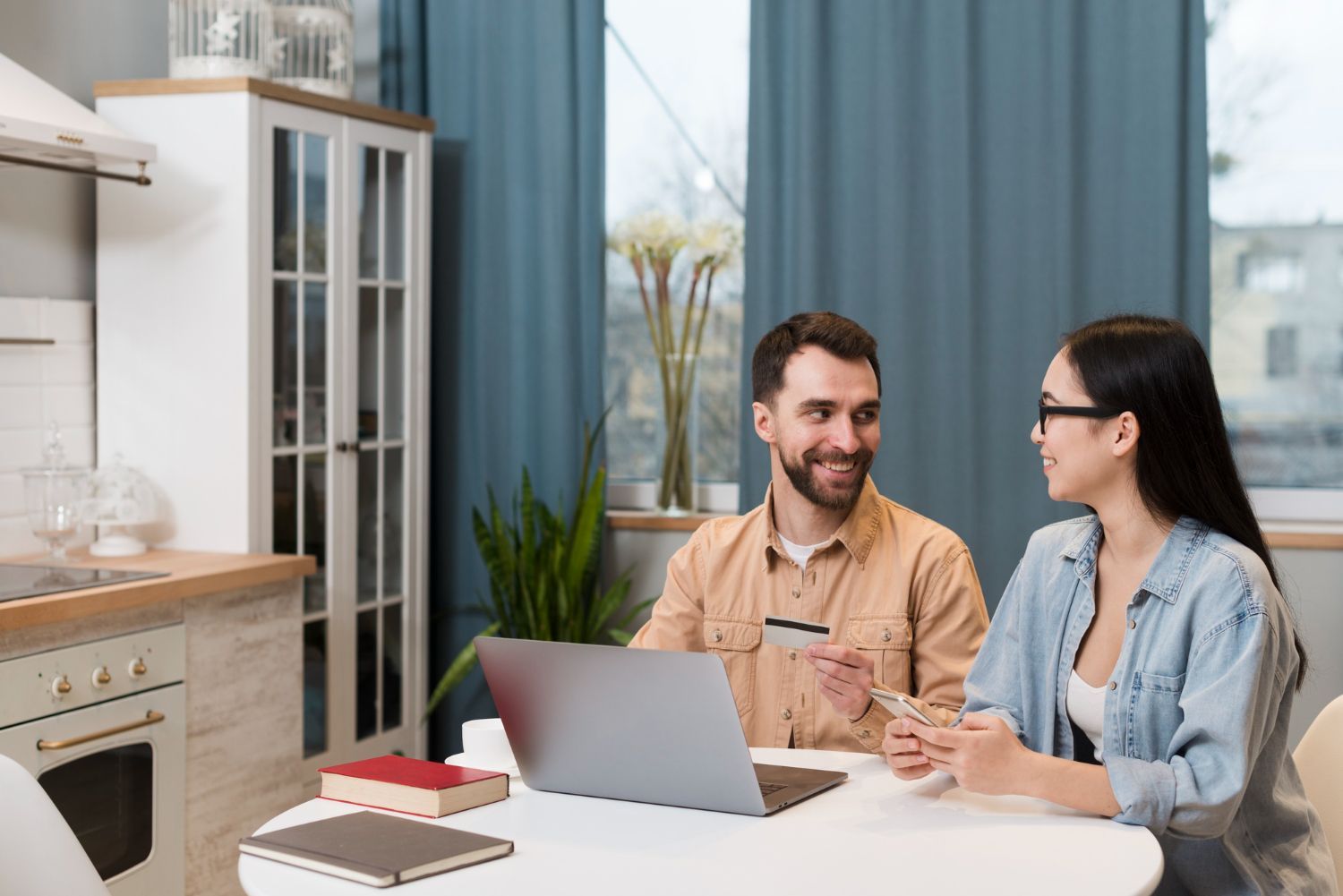 Couple in kitchen using laptop, man holding credit card, woman holding phone, both smiling.