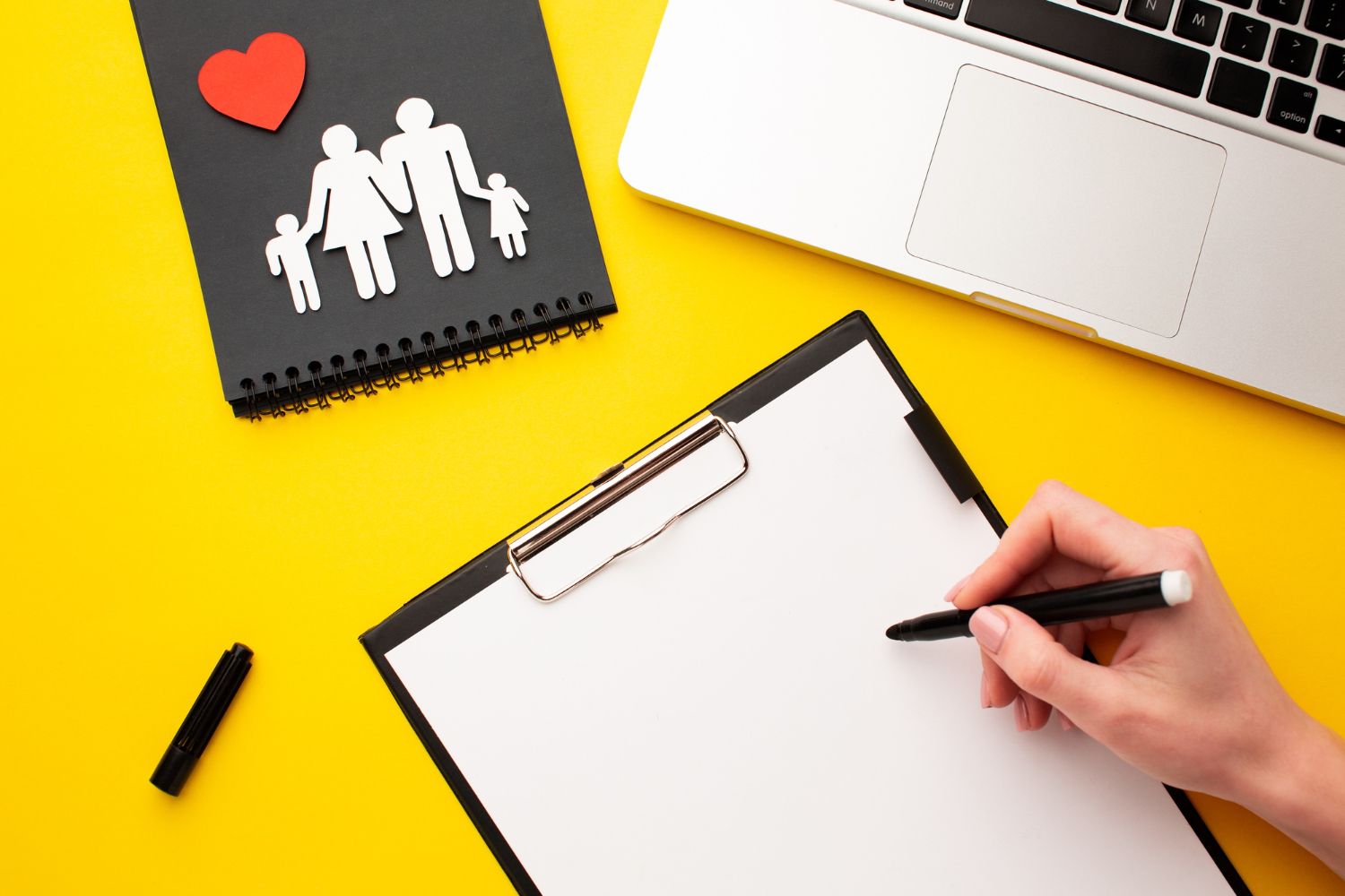 Yellow desk with notepad, family cutout, laptop, clipboard, and hand writing.