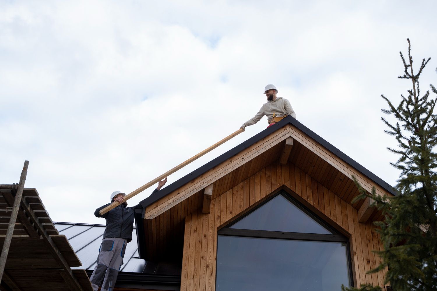 Two construction workers on a roof installing a long wooden beam on a building. Overcast sky.