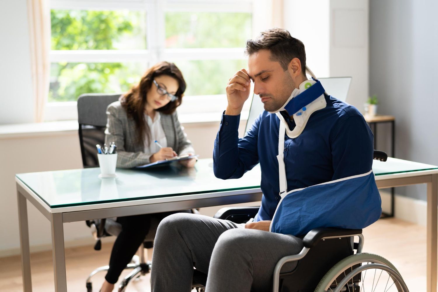 Man in wheelchair, sling, and neck brace meets with a woman at a desk. He looks distressed.