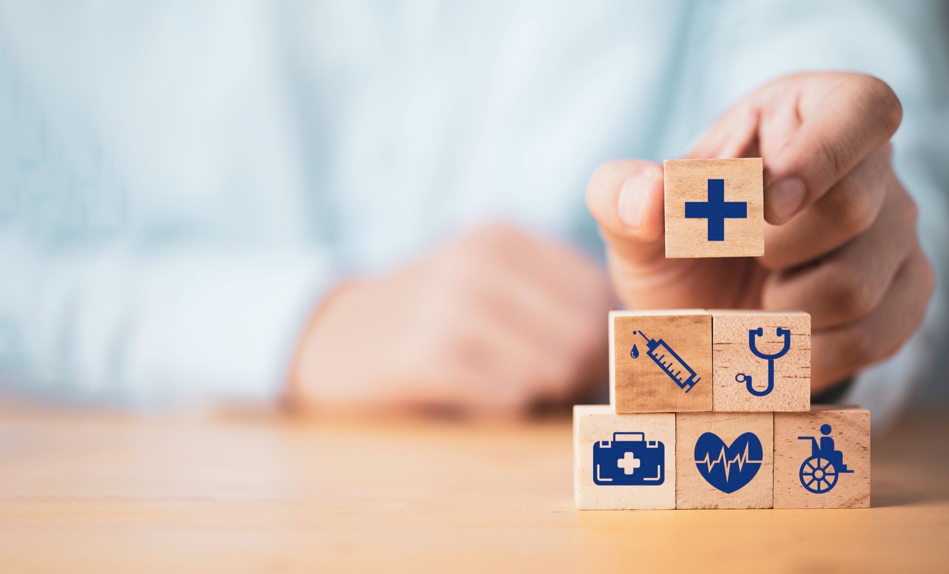 Person stacking wooden blocks with health-related symbols: cross, syringe, stethoscope, first aid kit, heart, wheelchair.