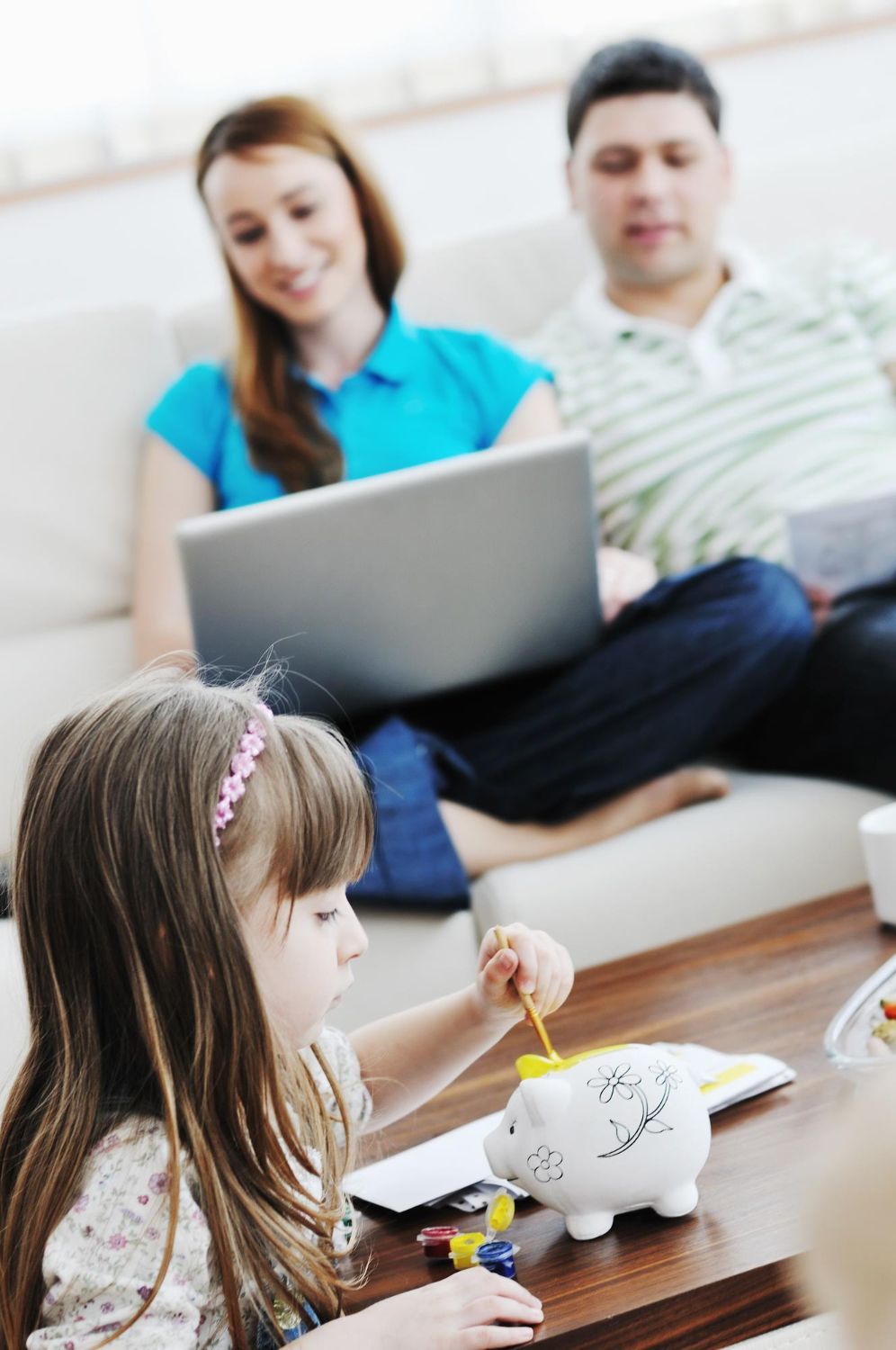 Girl putting money in piggy bank while parents work on laptop and look at documents.
