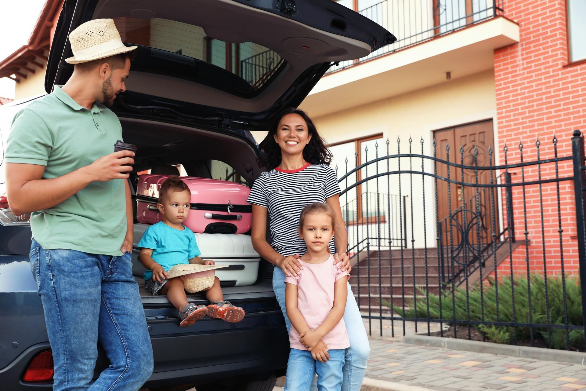 Family packing car for vacation; smiles at camera. Pink luggage, house in background.