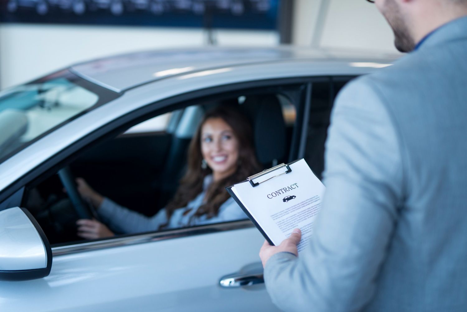 Car salesperson holds clipboard near driver inside a silver car; woman smiles, ready to sign.
