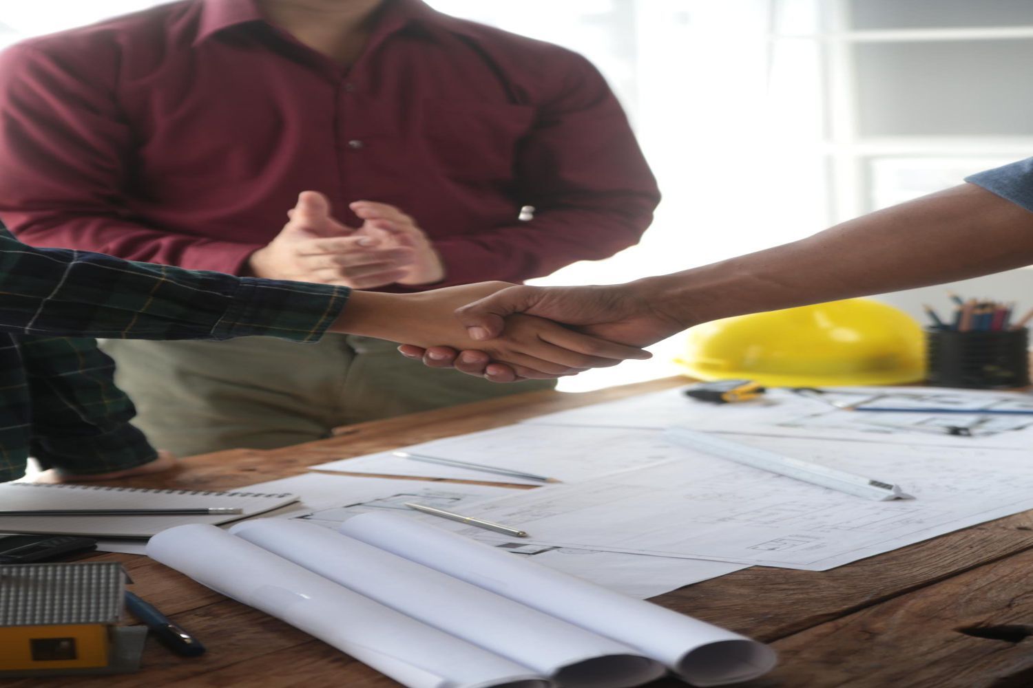Two people shake hands over blueprints at a table, another person in the background applauds.
