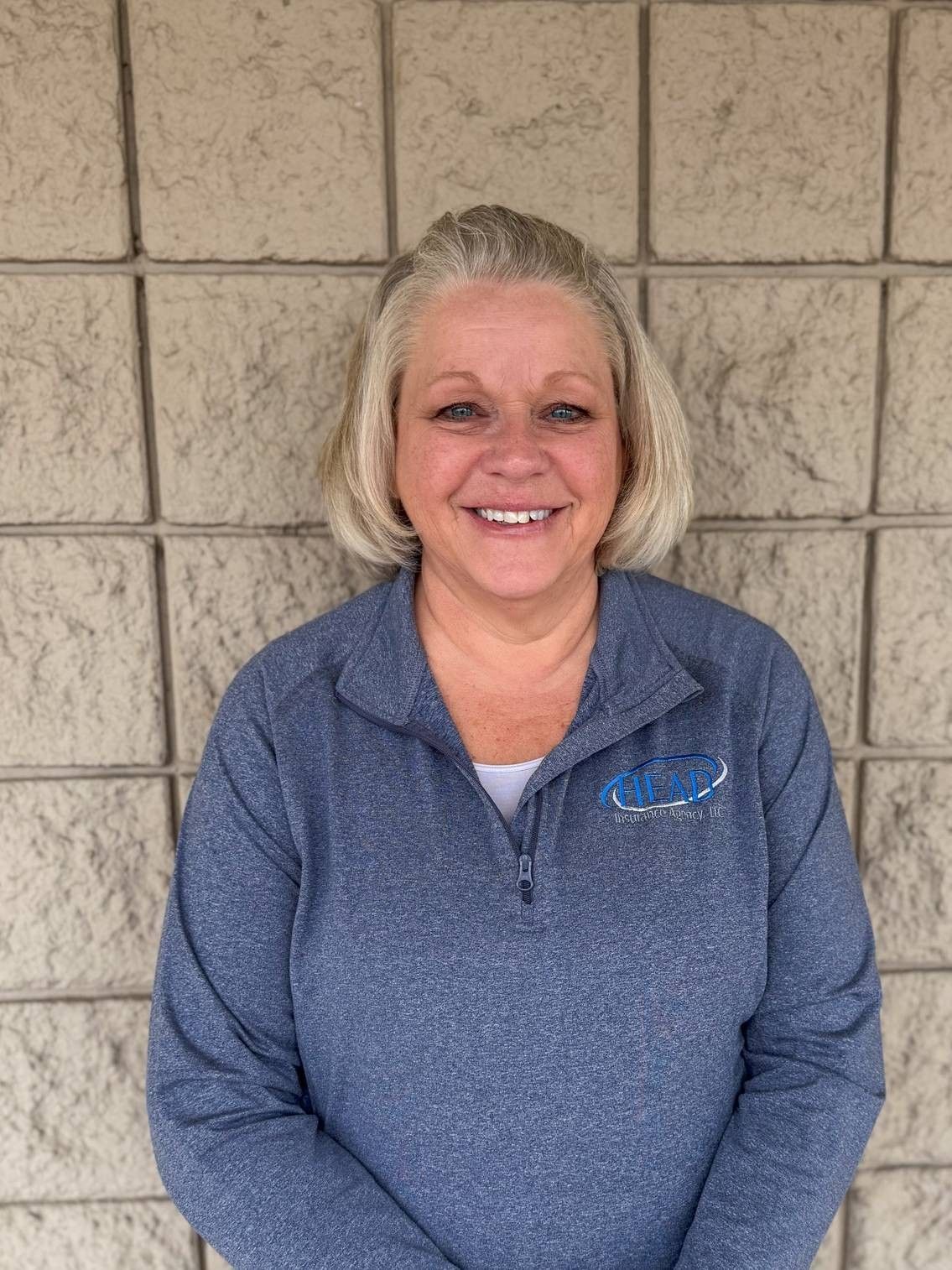 Woman with blonde hair smiling, wearing a floral navy top, leaning against a textured gray wall.