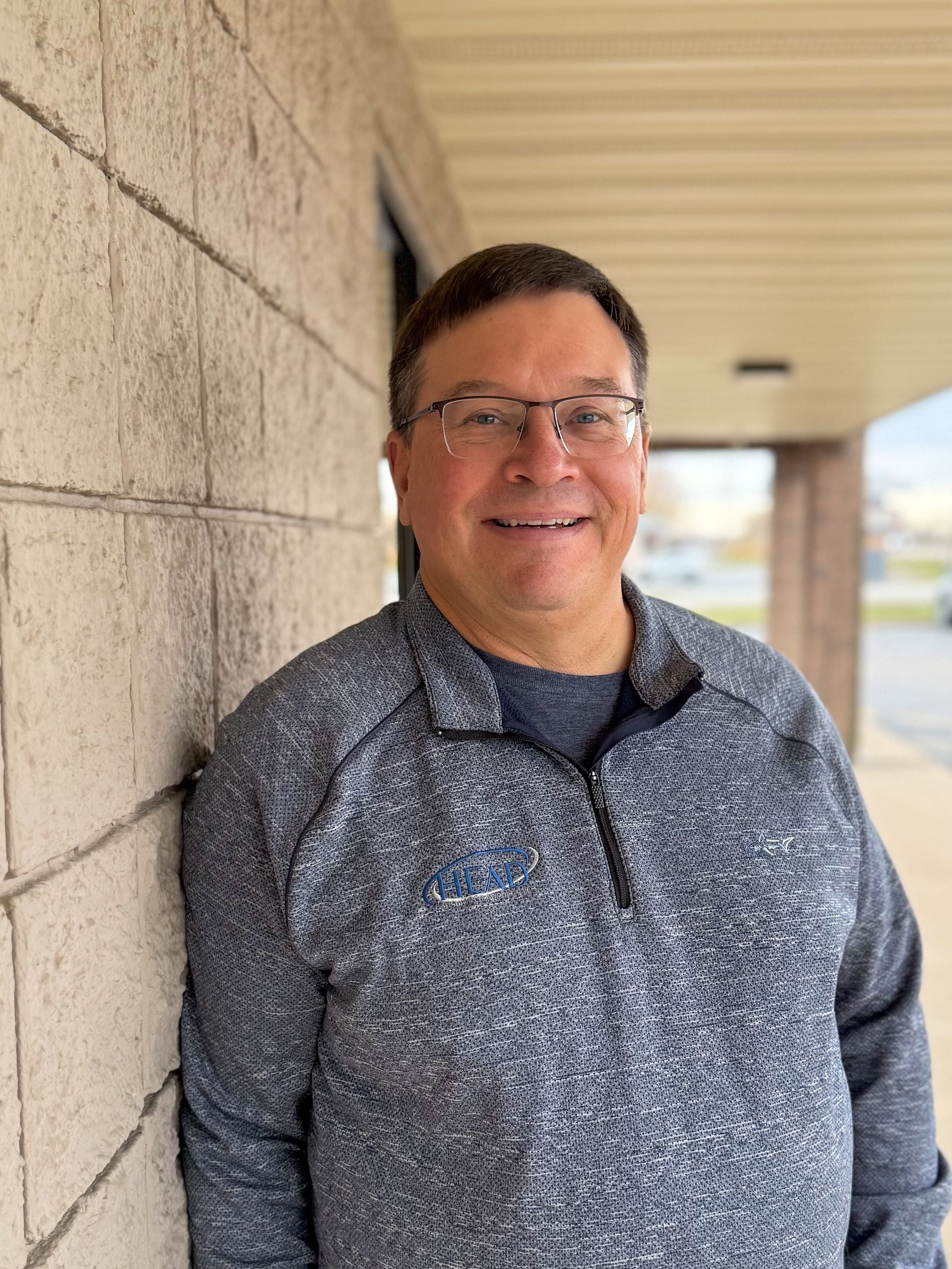 Man in blue polo shirt and glasses smiles in front of a textured wall.