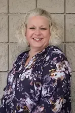 Woman with blonde hair smiling, wearing a floral navy top, leaning against a textured gray wall.