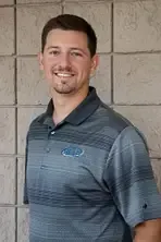 Man in gray polo shirt smiles, standing in front of a textured wall.