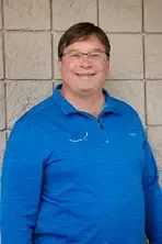 Man in blue polo shirt and glasses smiles in front of a textured wall.
