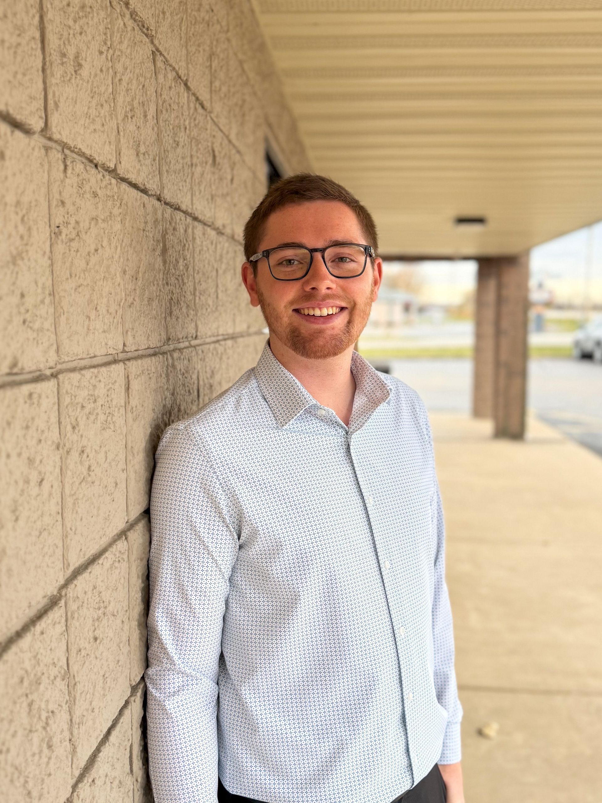 Man with glasses smiling, gray shirt, standing in front of a concrete block wall.