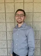 Man with glasses smiling, gray shirt, standing in front of a concrete block wall.