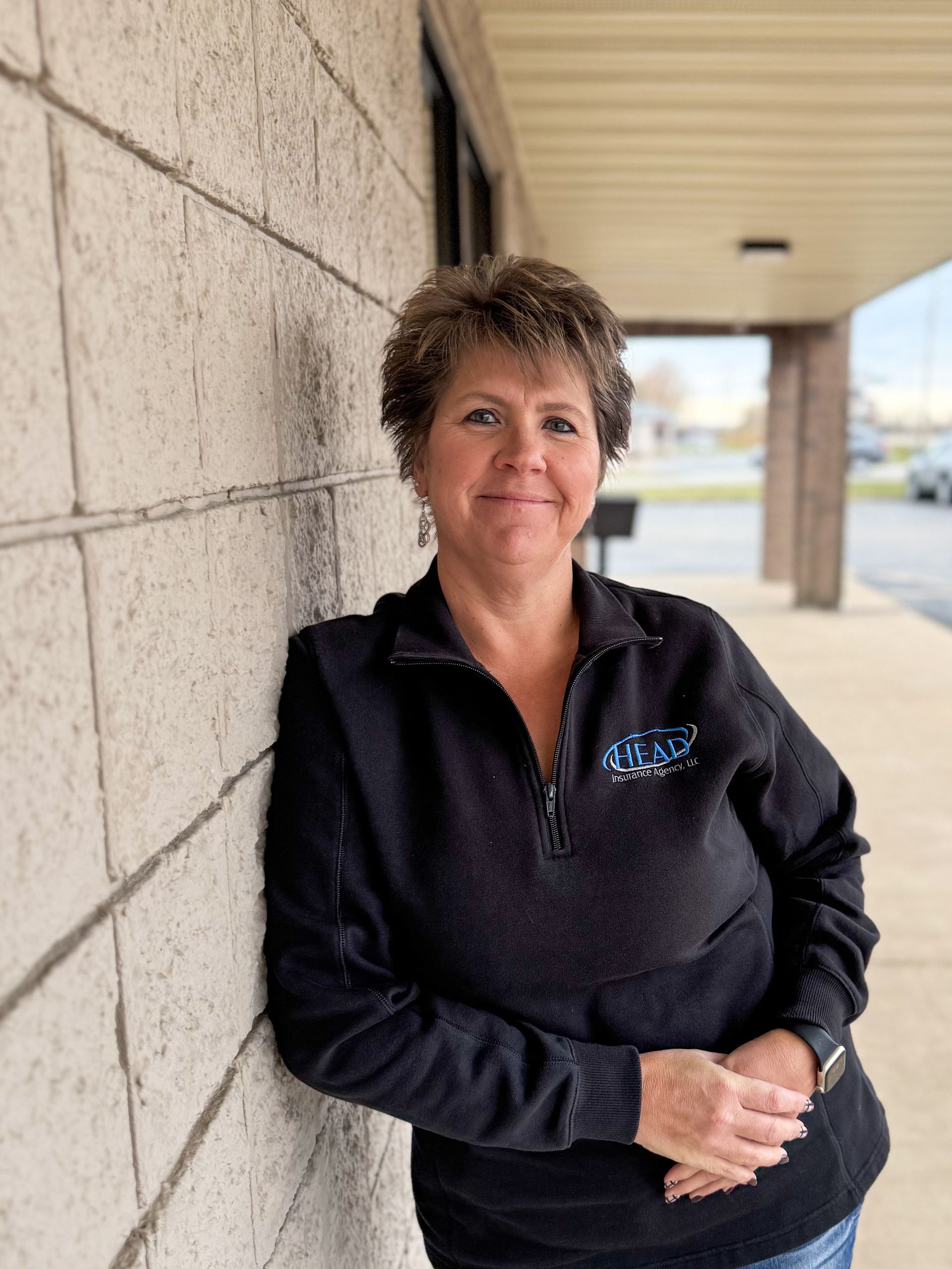 Woman smiling, leaning against a textured wall, wearing a black top and jeans.