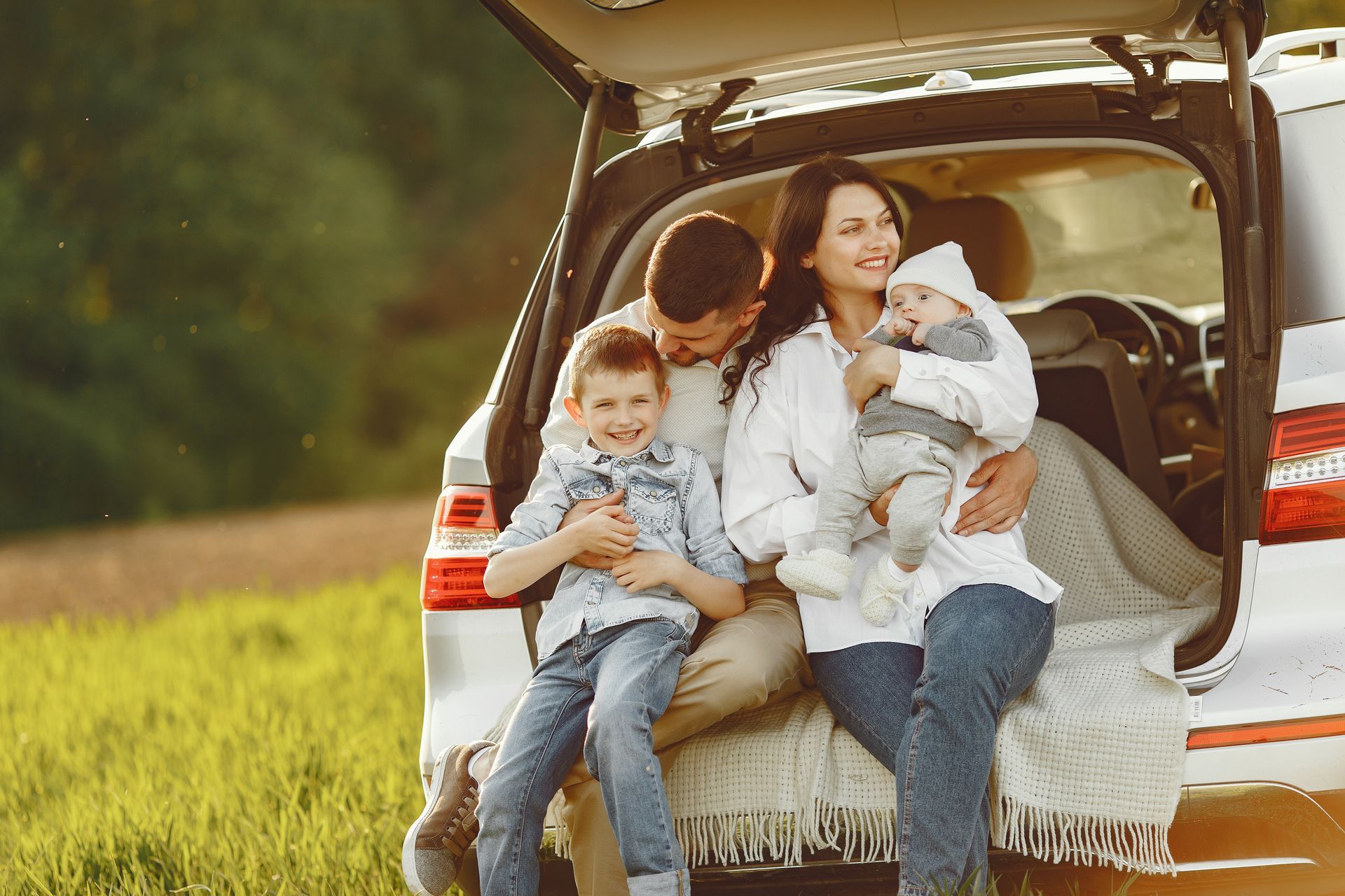 Family sits together in the open trunk of a car in a field; they are smiling.