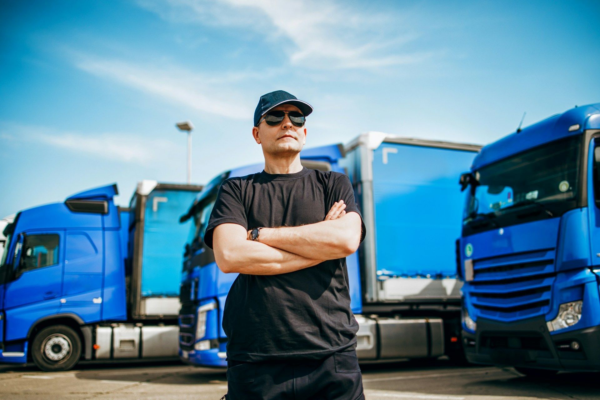 Man in sunglasses and hat with arms crossed, standing in front of several blue semi-trucks on a sunny day.