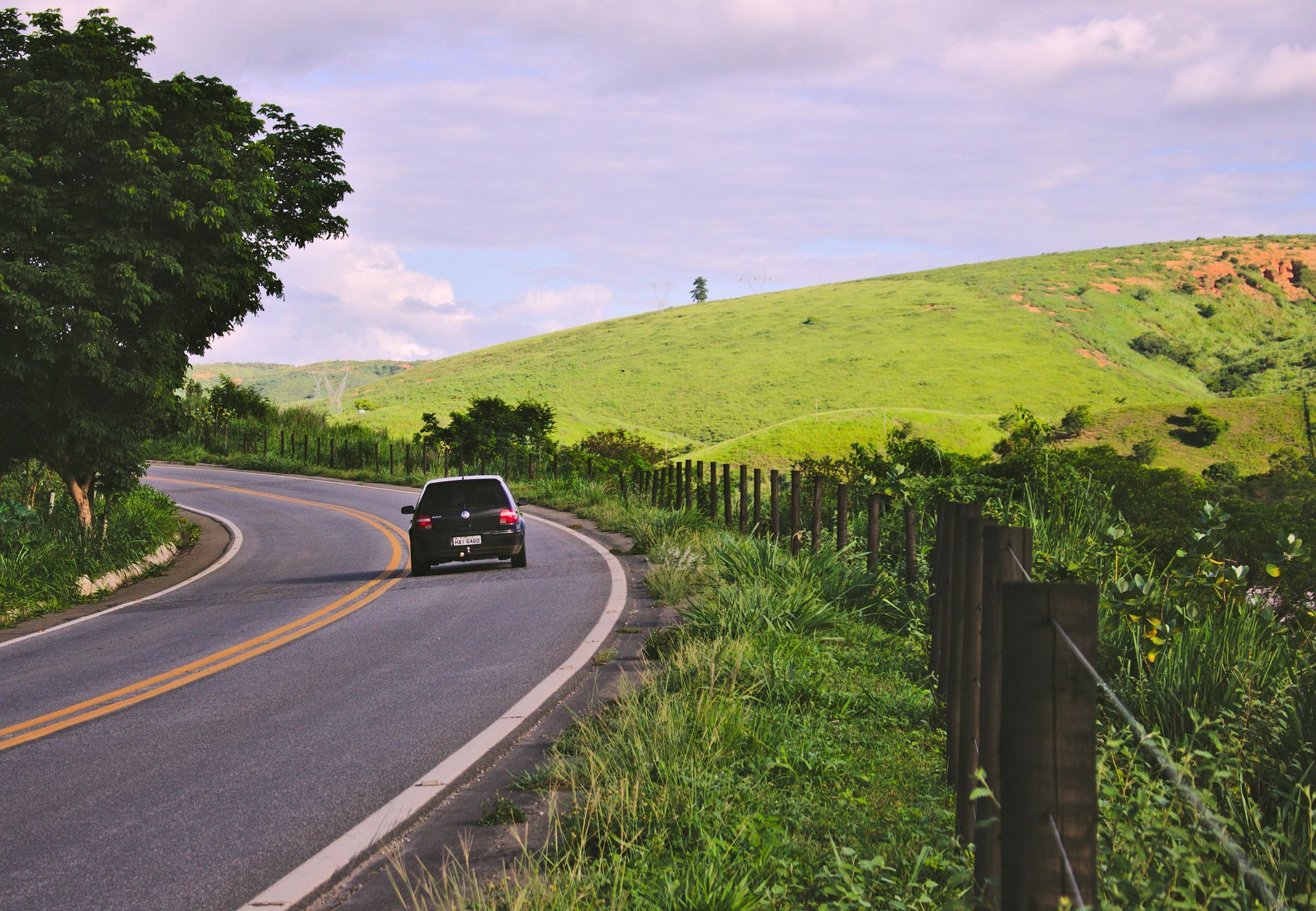 Black car on a winding road, surrounded by green hills and a wooden fence, on a sunny day.