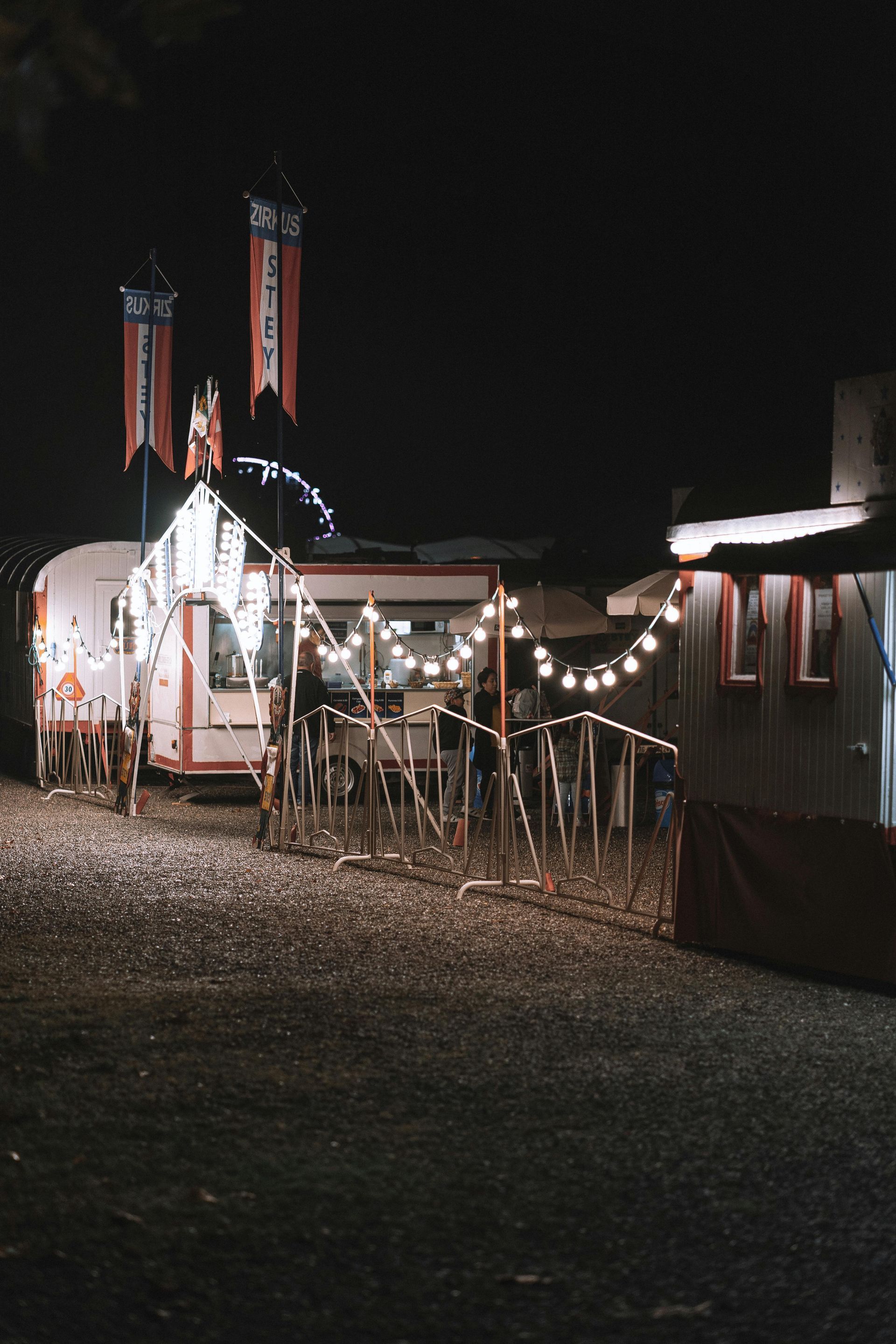Nighttime fairground with lit stalls, string lights, and flags.