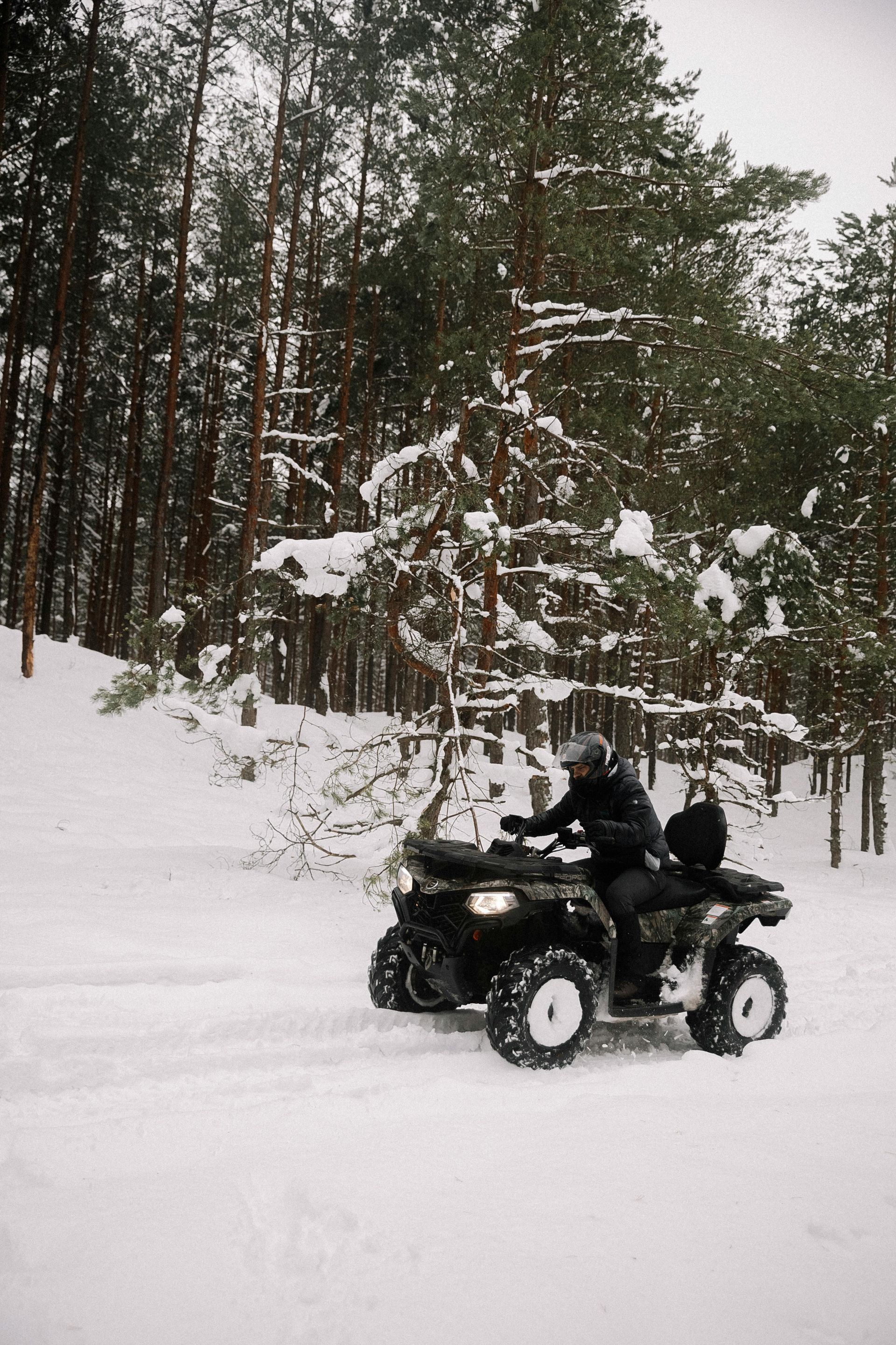 ATV in snowy forest, two people riding. Black ATV on a snow-covered path, trees in the background.