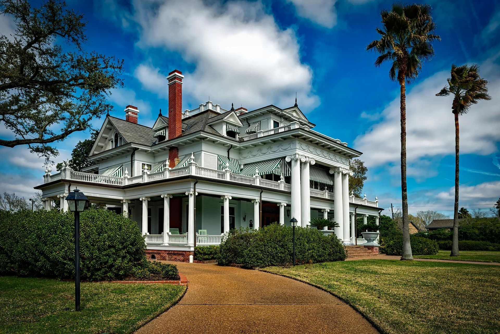 Elegant white Victorian house with porch and columns, brown driveway, palm trees, and blue sky.
