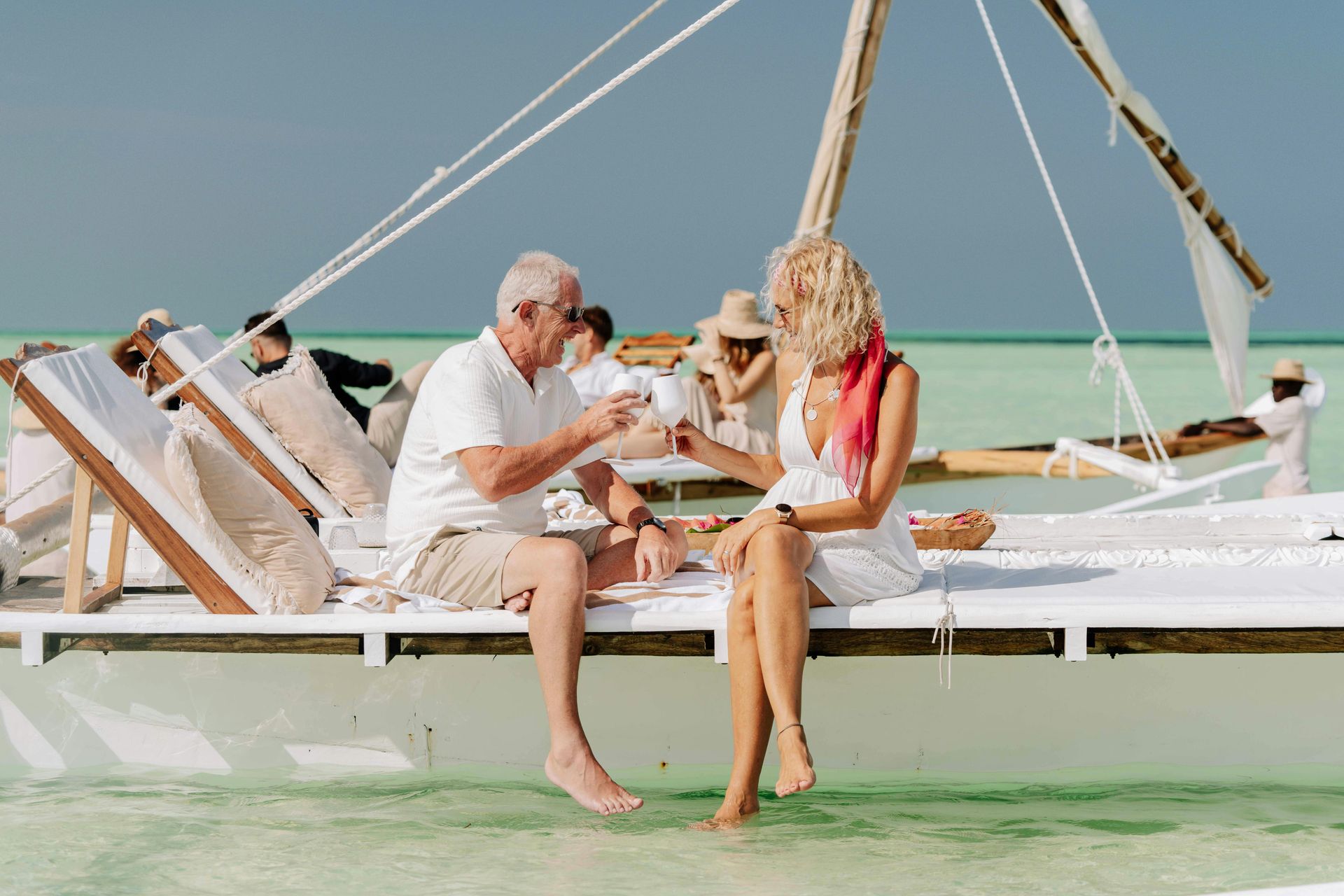 Couple toasting champagne on a white dock over turquoise water; other guests in the background.