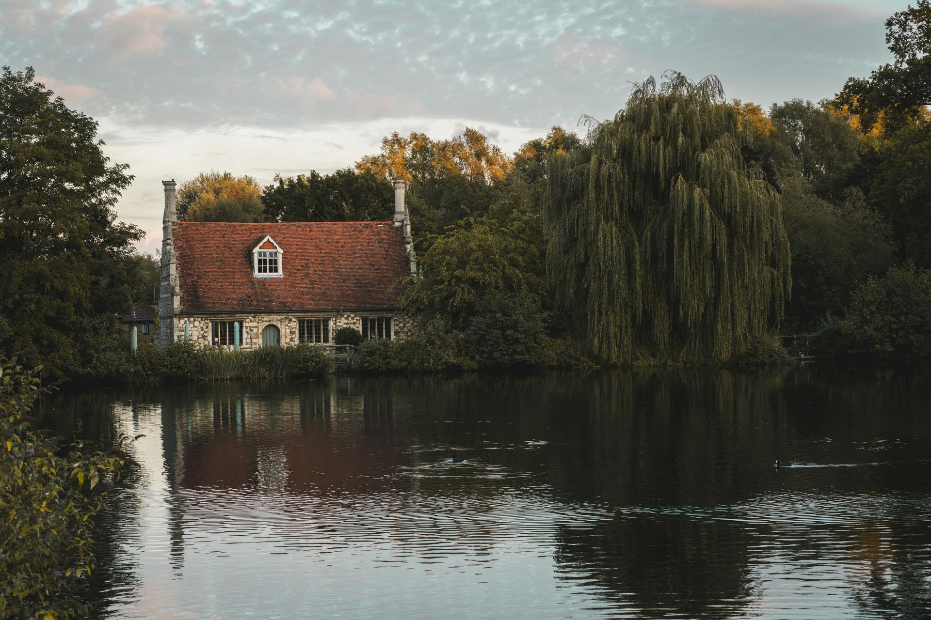 Cottage by water, red roof, stone walls, reflected in calm water, surrounded by trees. Cloudy sky.